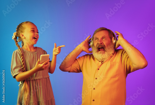 Crazy music of young generation. Senior man spending happy time with granddaughter in neon. Joyful elderly lifestyle, family, childhood, tech concept. Using headphones, smartphone. Copyspace.