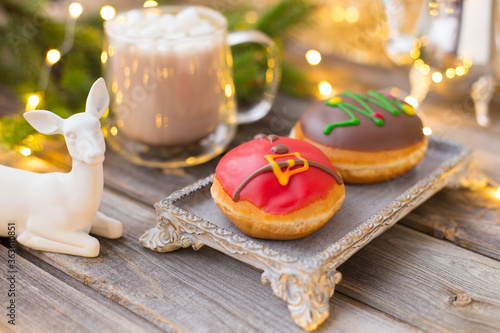 Christmas chocolate donuts with Santa Claus icing and new year tree on ceramic stand. Glass cup of cocoa with marshmallows on wooden table. Spruce branch, present, cone, sugar bowl,white deer,garland