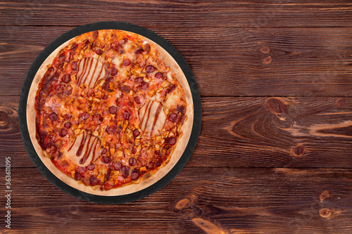 Delicious BBQ pizza with ham, bbq sauce, bacon and salami on a round slate platter which is on wooden table, top view and copy space