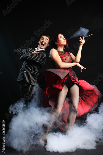 Fototapeta Naklejka Na Ścianę i Meble -  Funny man with mustache in suit with sexy woman in luxury evening dress who smoking mouthpiece and around many smoke isolated on black background