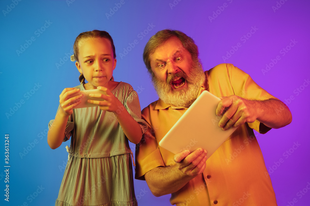 Playing videogames, crazy excited. Senior man spending happy time with granddaughter in neon. Joyful elderly lifestyle, family, childhood, tech concept. Playing with smartphone, tablet. Copyspace.