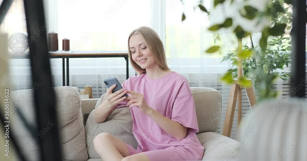 Happy woman in pajamas text messaging using mobile phone on sofa at home Happy woman in pajamas text messaging using mobile phone on sofa at home
