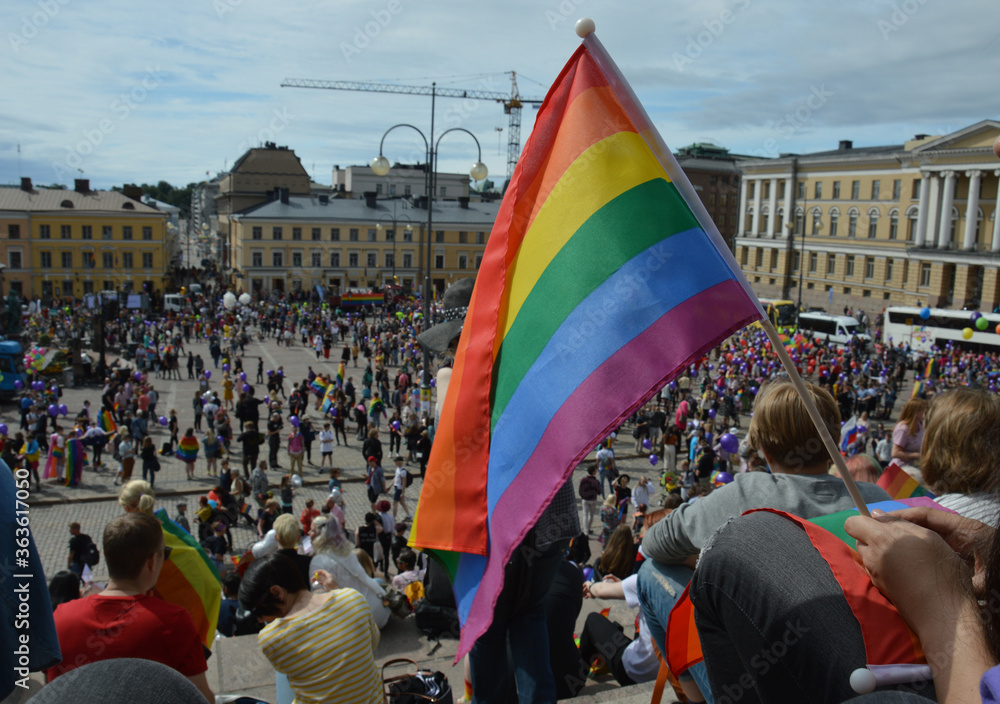 Helsinki Pride 2019 - crowd gathered on Senate Square to support diversity and inclusion. Hand is holding a rainbow flag in the foreground. Selective focus.