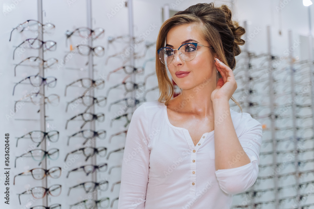 Fototapeta premium Happy female client or optician is standing with set of glasses in background in optical shop. Woman in spectacles. Eyesight correction.