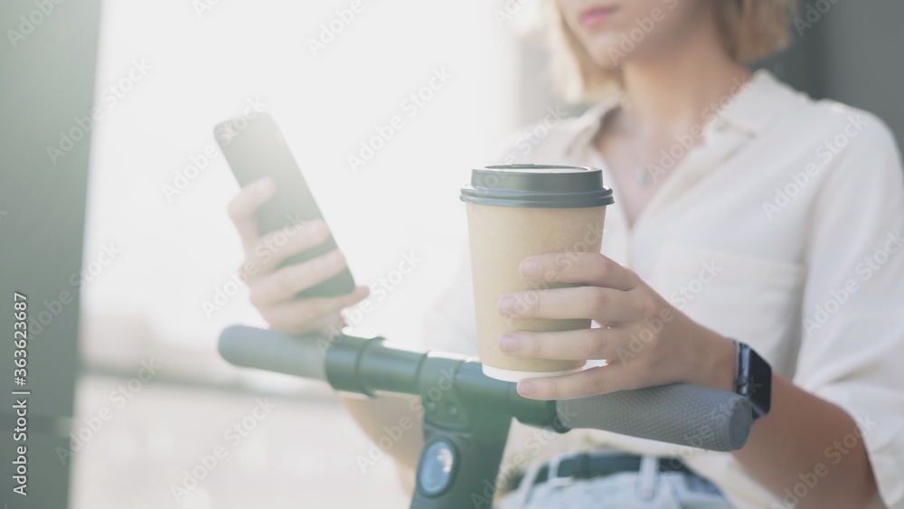 Woman is using smartphone and drinking coffee after ride on electric kick scooter Woman is using smartphone and drinking coffee after ride on electric kick scooter