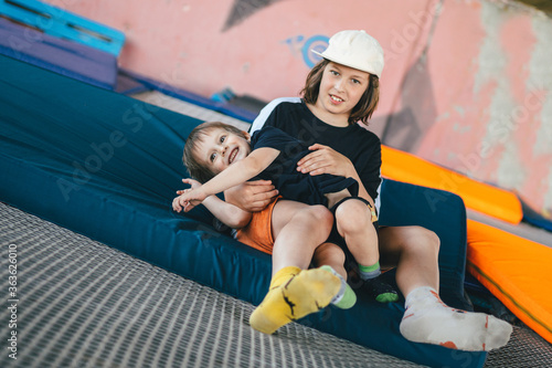 Two happy emotions children, brothers playing and having fun while jumping on bouncing trampoline in playground in summer. Hug, friendship and childhood concept. Active lifestyle of children
