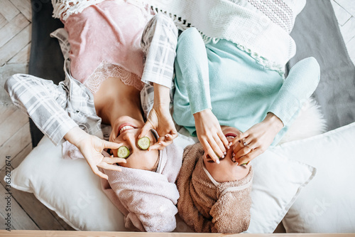 Two girls make homemade face and hair beauty masks. Cucumbers for the freshness of the skin around the eyes. Women take care of youthful skin. Girlfriends laugh at home lying on the floor on pillows.