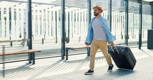Caucasian young stylish man traveller in hat and with backpack walking at bus station and carrying suitcase on wheels. Handsome male tourist strolling outdoor from train station or aeroport. photo