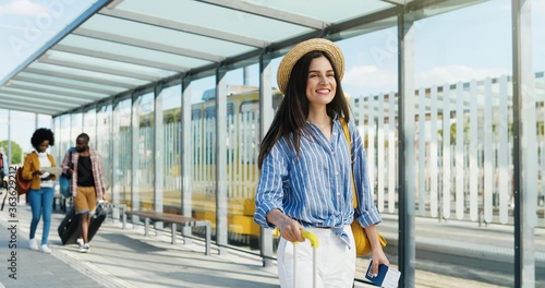 Caucasian young beautiful woman in hat walking outdoor and carrying suitcase on wheels. Pretty female tourist strolling at bus and tram stop and smiling. saying hello or hi to handsome guy.