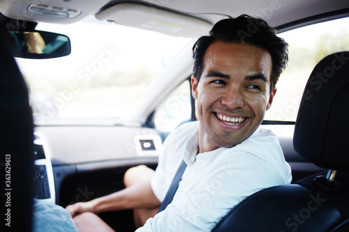 Young Latin man with bright smile laughs to his friends sitting behind him in the car during their joint road trip, happy cheerful male talking with passengers while sitting in comfortable automobile