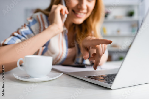 Cropped view of smiling woman pointing with finger at laptop while talking on smartphone near cup of coffee on table © LIGHTFIELD STUDIOS