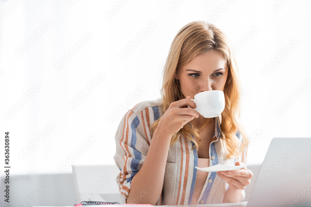 Selective focus of pretty woman drinking coffee while working with laptop Selective focus of pretty woman drinking coffee while working with laptop