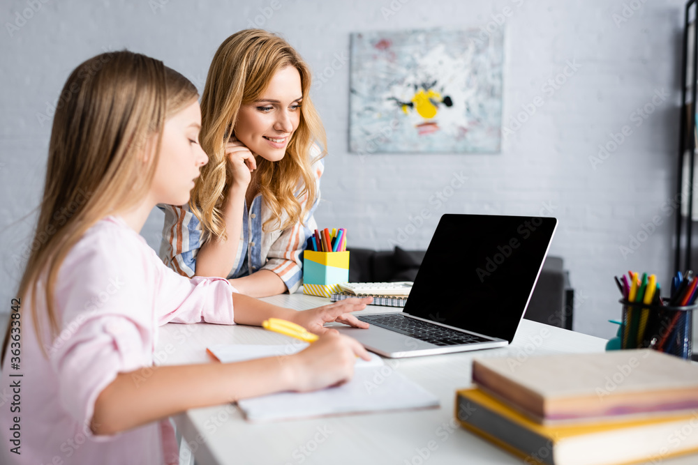 Selective focus of smiling woman standing near kid using laptop during electronic learning at home Selective focus of smiling woman standing near kid using laptop during electronic learning at home