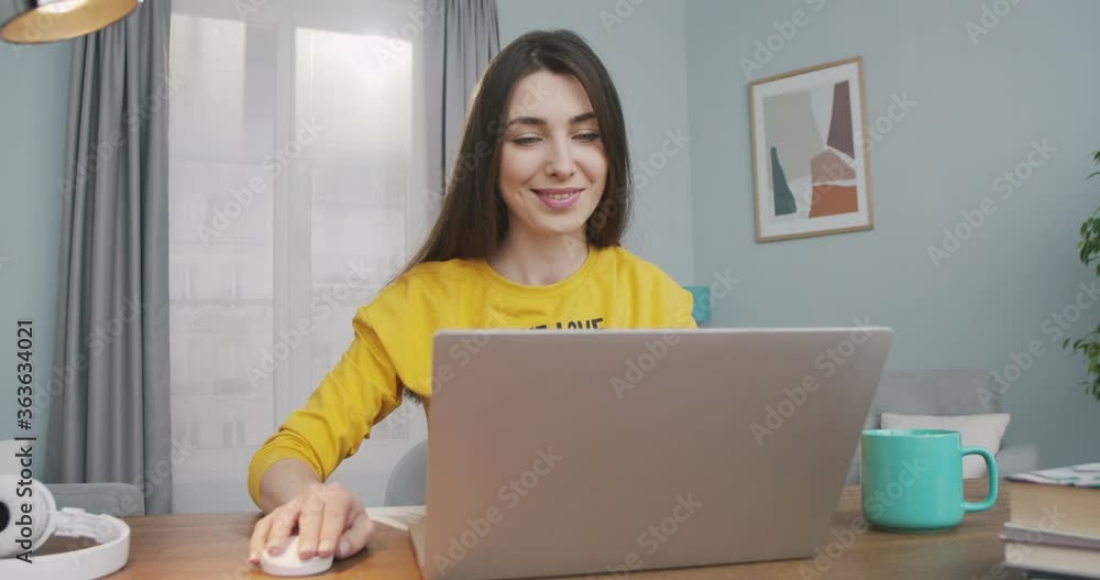 Cheerful beautiful girl opens computer and typing while working at home in good mood. Pretty Caucasian joyful young female browsing on laptop while sitting at table in room. Freelancer concept Cheerful beautiful girl opens computer and typing while working at home in good mood. Pretty Caucasian joyful young female browsing on laptop while sitting at table in room. Freelancer concept