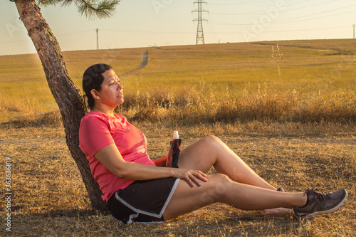 Young athletic woman resting on a tree after running wearing sports clothing, striped shirt and black skirt with a beautiful yellow field during a sunset while drinking water from a black bottle