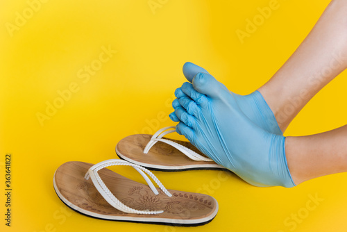 Legs of a girl dressed in rubber blue medical sterile gloves, shod in white flip flops on an orange yellow plain background. A joke of a safe rest on the sea in the sand, protection against a virus