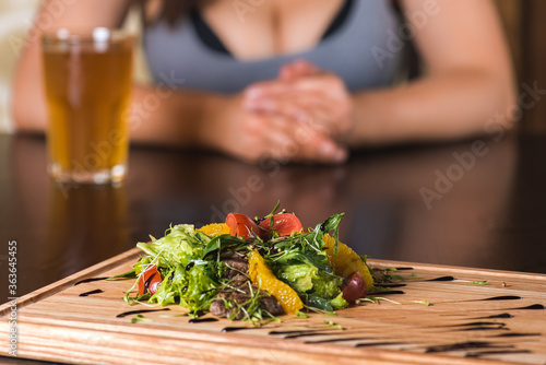 girl sitting at the table in front of her salad of greens, orange, tomato, veal, fresh vegetables on a wooden tray, blackboard, dish. Horizontal photo