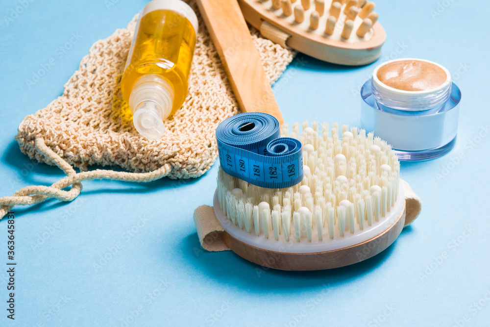 body oil, measuring tape, knitted washcloth, natural homemade scrub in a jar and brush for dry massage on a blue background, a set for anti-cellulite massage body oil, measuring tape, knitted washcloth, natural homemade scrub in a jar and brush for dry massage on a blue background, a set for anti-cellulite massage