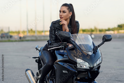 Attractive girl with long hair in black leather jacket and pants on outdoors parking with stylish sports motorcycle at sunset. © gorynvd