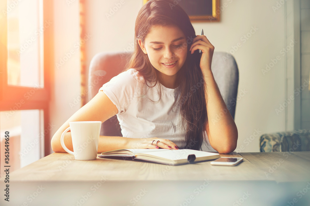 Smiling intelligent young woman with gorgeous brunette hair reading interesting book sitting at table with mug of tea in coworking space during leisure time.Cheerful skilled student preparing for exam Smiling intelligent young woman with gorgeous brunette hair reading interesting book sitting at table with mug of tea in coworking space during leisure time.Cheerful skilled student preparing for exam