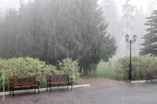 Rain and hail on a summer day. Large puddles on the sidewalk during heavy rain. Heavy rain with snow and hail in the Park. Wet benches on the paved path. Spray from drops and bubbles in large puddles.