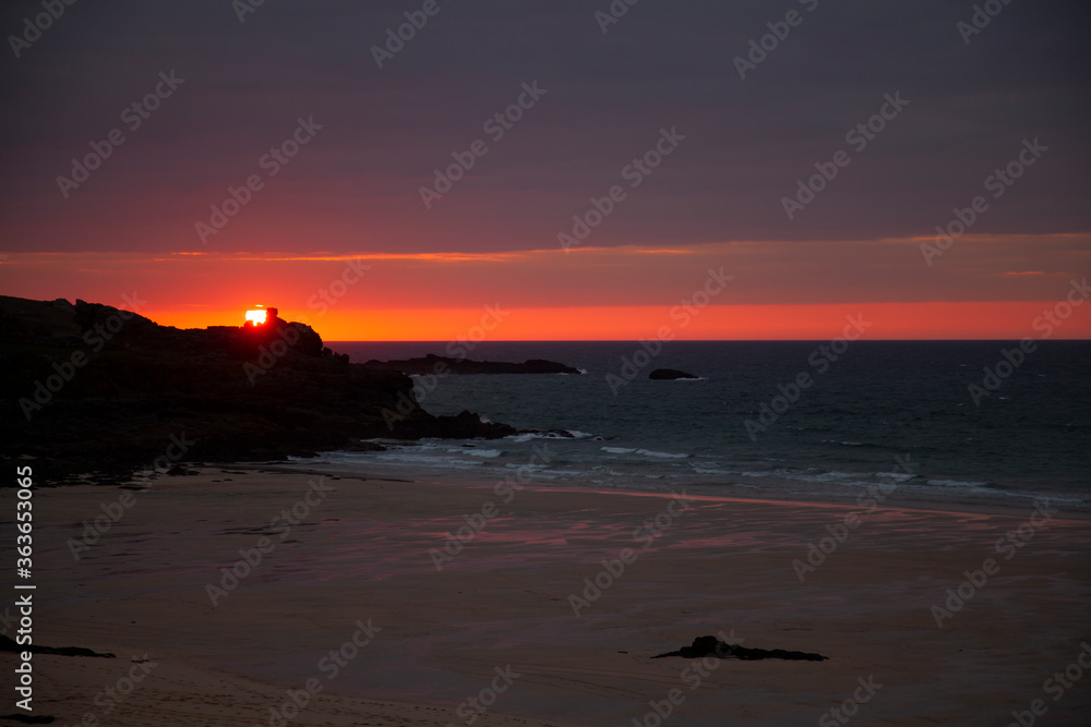 Image of a sunset over Atlantic ocean as photographed from the rugged coast of Cornwall near St. Ives. The photo shows a bay, sandy beach, calm sea, cloudy sky, silhouettes of rocks and the horizon.