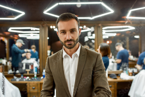 People at barbershop. Young handsome bearded man wearing white shirt and jacket looking at camera, visiting barber shop or beauty salon