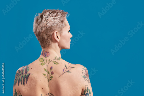 Beauty portrait of a young attractive half naked tattooed woman with perfect skin looking away, posing back to camera isolated over blue background