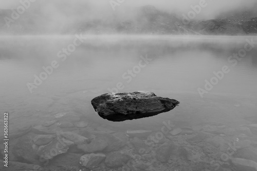 La brume et les nuages tombent sur un petit lac dans les montagnes des Pyrénées françaises. Un rocher émerge de l'eau. La photo est en noir et blanc, le paysage est calme, reposant et  paisible. photo