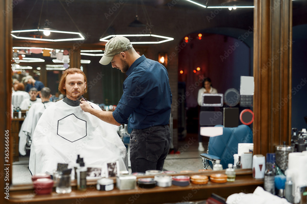 Obraz premium Grooming. Young professional barber trimming beard of a young handsome redhead guy sitting in barbershop chair. Focus on a barber and client