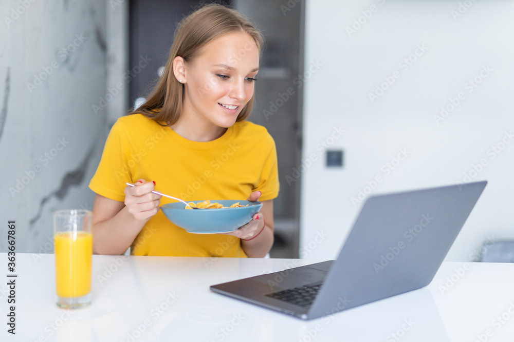 Beautiful young woman eat breakfast leaning on the kitchen table with laptop. Beautiful young woman eat breakfast leaning on the kitchen table with laptop.