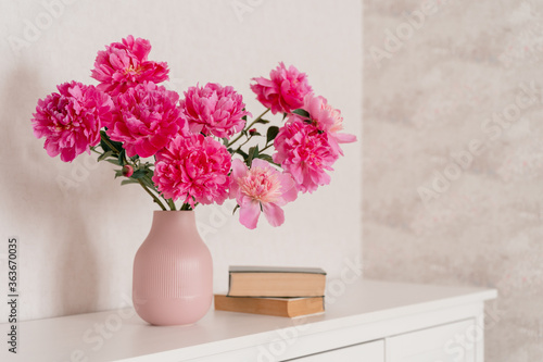 Fototapeta Naklejka Na Ścianę i Meble -  big bouquet of  peonies in a pink vase in a bright room are on a white chest of drawers, books are nearby. cozy house, fresh flowers at home, home greenhouse, minimal design, loft house