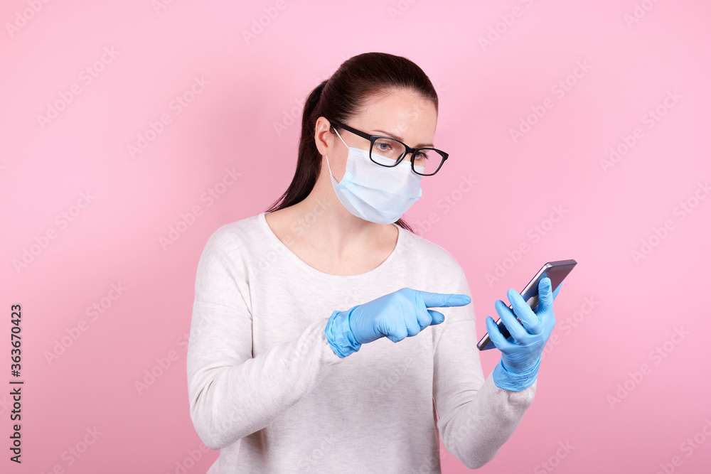 Portrait of a brunette girl with eyeglasses in a medical mask and blue rubber gloves using cellphone. Isolated on pink background.