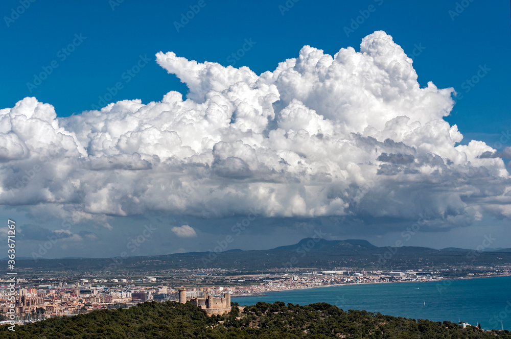 Landscape with cumulus type clouds, trying to evolve into cumulonimbus and preparing rainfall, on a bay with sea and the city of Palma, with a circular castle on top of a small hill in the foreground