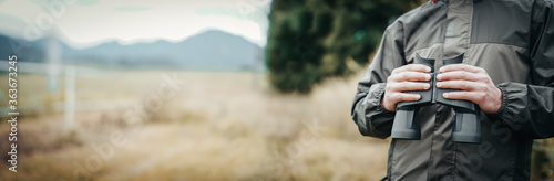 Panorama close up of a man dressed in a hunter jacket holds binoculars in his hand.