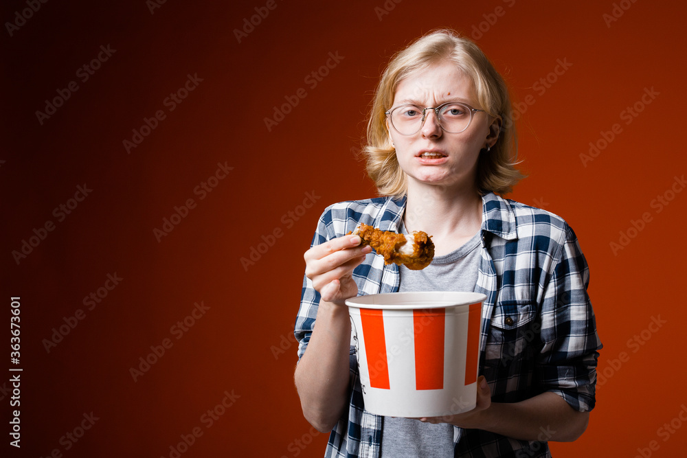 A dissatisfied blonde with glasses holds a bucket of chicken and looks at the camera grimacing against a red background. A dissatisfied blonde with glasses holds a bucket of chicken and looks at the camera grimacing against a red background.