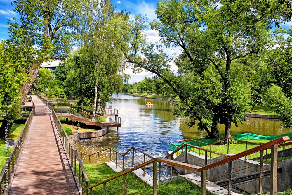 city park landscape at daytime with wooden pathway along pond embankment against blue sky with clouds background. Summer in Moscow city Russia city park landscape at daytime with wooden pathway along pond embankment against blue sky with clouds background. Summer in Moscow city Russia