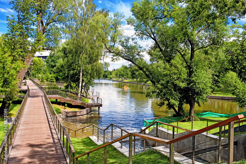 city park landscape at daytime with wooden pathway along pond embankment against blue sky with clouds background. Summer in Moscow city Russia