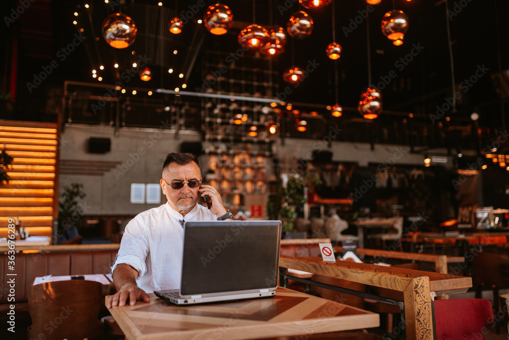 A middle-aged caucasian businessman with glasses in a white shirt is sitting in a cafe, talking on the phone and working on a laptop A middle-aged caucasian businessman with glasses in a white shirt is sitting in a cafe, talking on the phone and working on a laptop