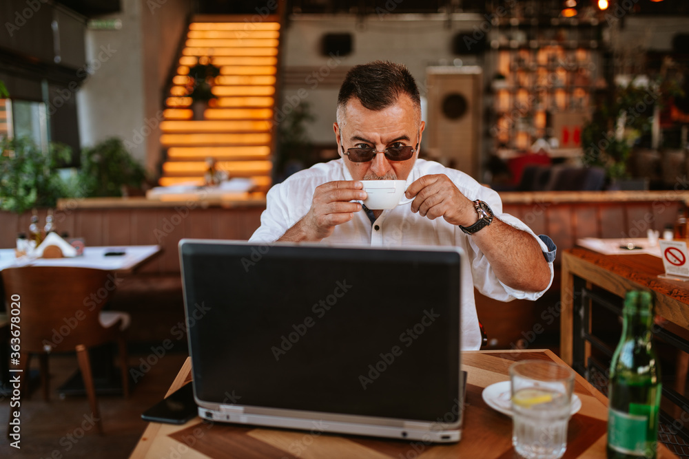 A middle-aged caucasian 
 casual businessman with glasses in a white shirt is sitting in a modern cafe, working on a laptop, drinking espresso coffee