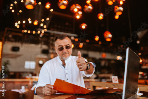 A middle-aged caucasian businessman with glasses in a white shirt is sitting in a cafe, giving thumbs up, working on a laptop and holding a business folder in his hand