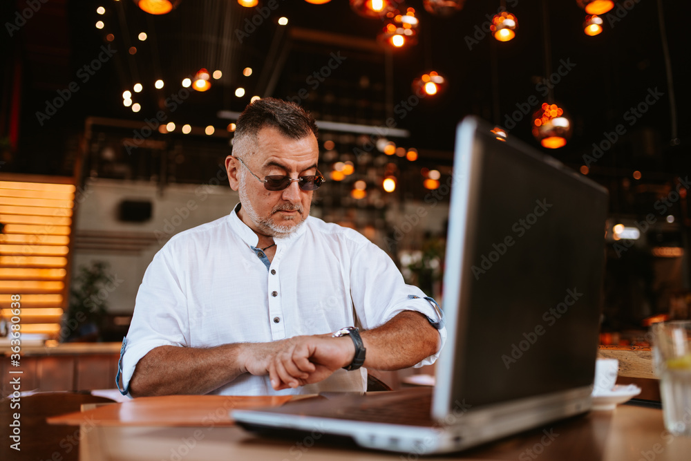A middle-aged caucasian man with glasses in a white shirt sitting in a cafe looking at his watch while a laptop is on the table A middle-aged caucasian man with glasses in a white shirt sitting in a cafe looking at his watch while a laptop is on the table