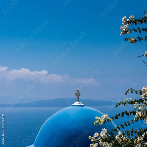 Detail of a blue church roof in Santorini island, Greece.. Bougainvillea white flowers on the foreground. Blue sea and sky in the background. Square format