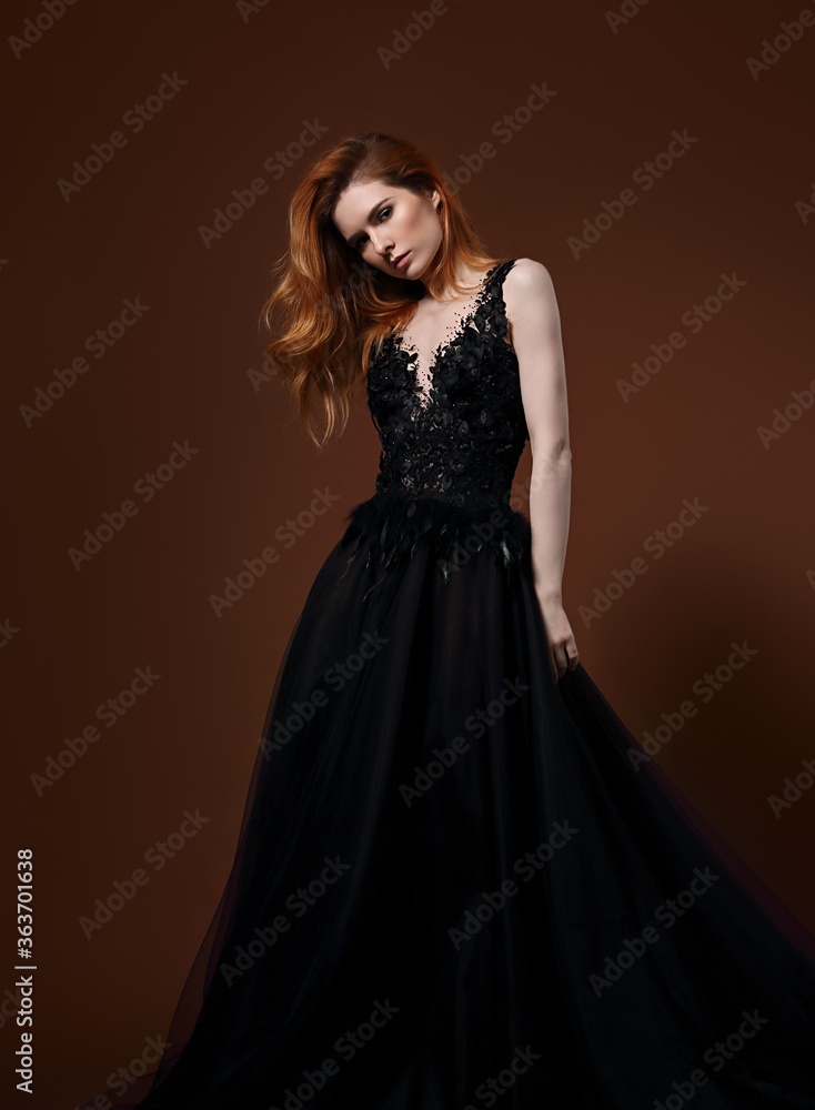 Beautiful elegant young model with bright redhead hairstyle posing in fashion chic black wedding dress with long skirt on studio bright orange background. Portrait Beautiful elegant young model with bright redhead hairstyle posing in fashion chic black wedding dress with long skirt on studio bright orange background. Portrait