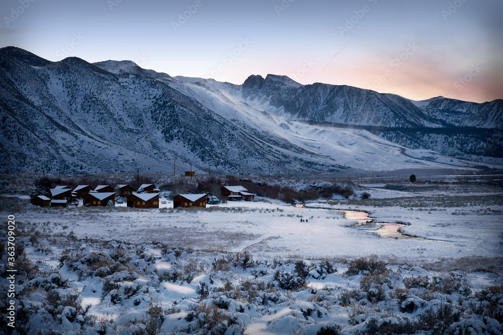 Snow covered mountains with small cabins nestled in the frozen valley next to a river reflecting the sunset