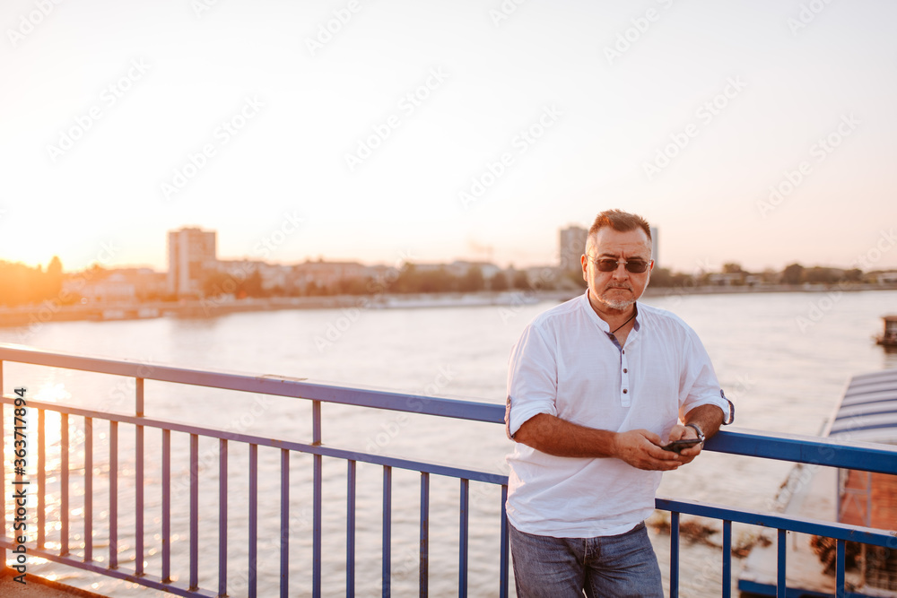 A middle-aged caucasian businessman with glasses, in a white shirt, is typing a message on the phone leaning against the bridge railing.