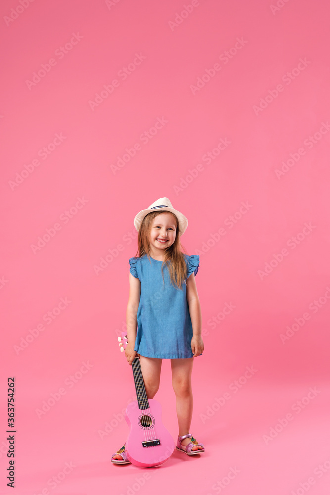 cheerful little girl in a hat poses with a ukulele guitar on a pink background. cheerful little girl in a hat poses with a ukulele guitar on a pink background.