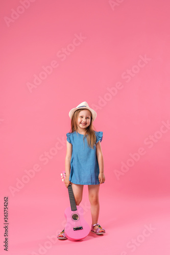 cheerful little girl in a hat poses with a ukulele guitar on a pink background.