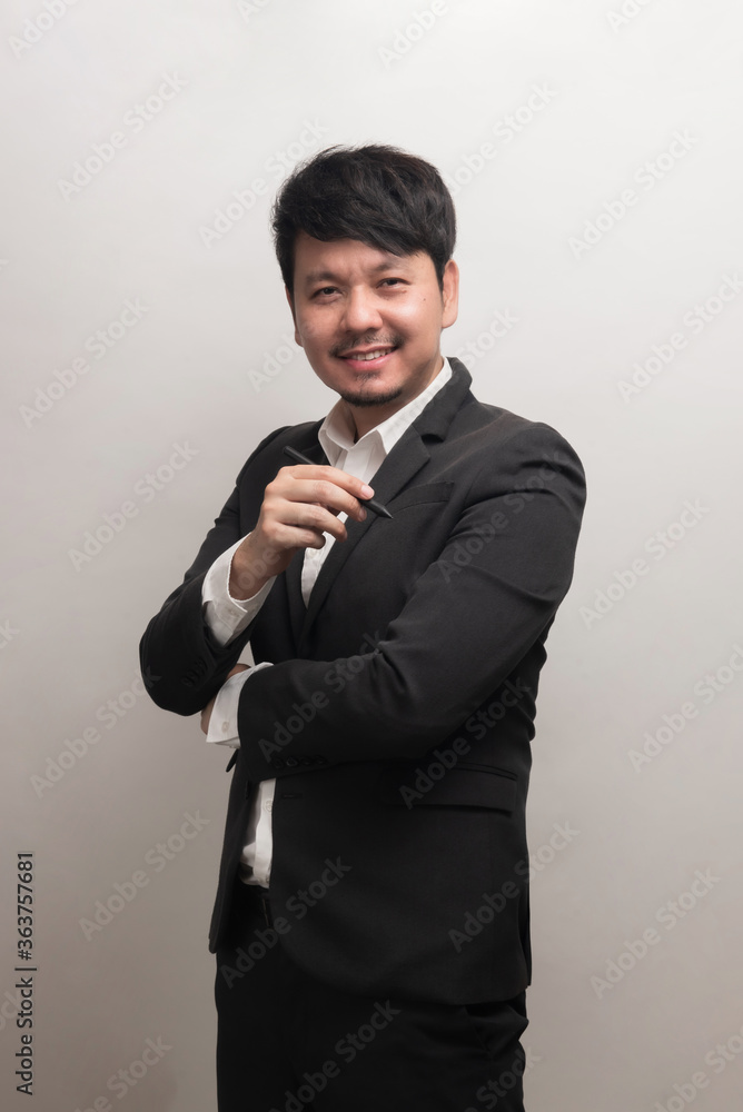 Smiling Asian businessman with white shirt and black suite holding pen, look at camera, isolate on grey background Smiling Asian businessman with white shirt and black suite holding pen, look at camera, isolate on grey background