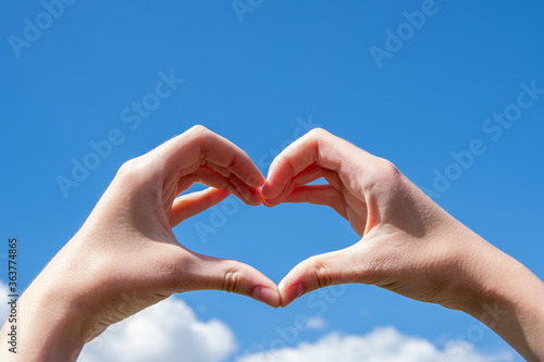 Close-up of hands making a heart symbol on a background of blue sky and clouds. Love, charity, valentines day concept